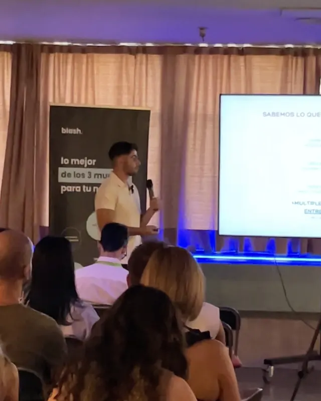 Alejandro dando una ponencia con micrófono junto a una pantalla, con el público sentado delante